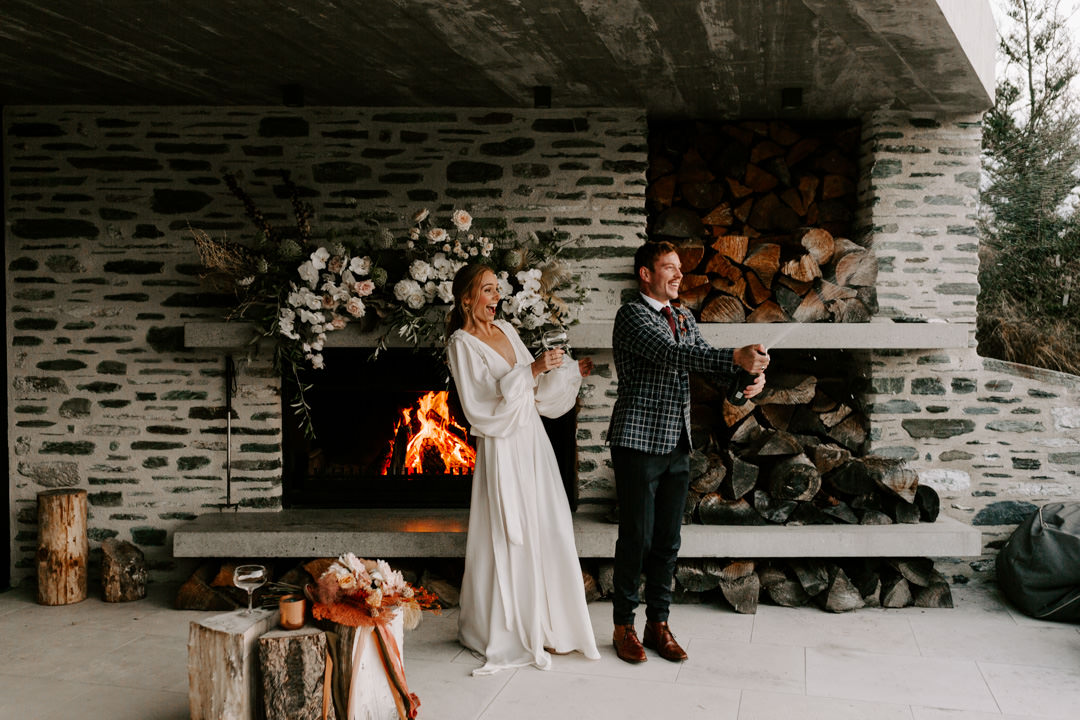 Fireplace and bride and groom popping champagne at Jacks Retreat Elopement Wedding in Queenstown, New Zealand by Dawn Thomson Photography